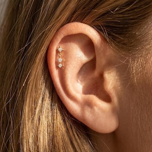 May include: A gold-colored ear piercing featuring a star-shaped stud and three dangling, round, clear gemstones. The jewelry is worn on the upper ear cartilage. The background shows light brown hair.