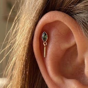 May include: A gold-colored earring with a dark green, oval-shaped gemstone and a dangling vertical bar. The earring is set in a person's ear, with the focus on the jewelry's design and details.