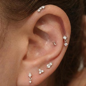 May include: Close-up of an ear adorned with multiple silver earrings. The earrings include a curved bar of three small stones, a star stud, a dangling square and round stone earring, a teardrop-shaped earring, a cluster of three stones, and a small, irregular-shaped stone earring.