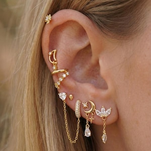 May include: An ear adorned with multiple gold earrings. The earrings include a star stud, a crescent moon and star dangle, a heart-shaped charm with a chain, and a cluster of small, sparkling stones. The jewelry is set against a blurred background.