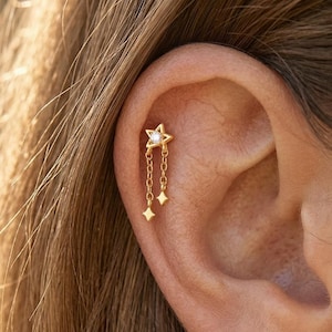 May include: Gold-colored ear piercing featuring a star design with a small, clear gem in the center. Two dangling star-shaped charms hang from delicate chains. The earring is shown on an ear.