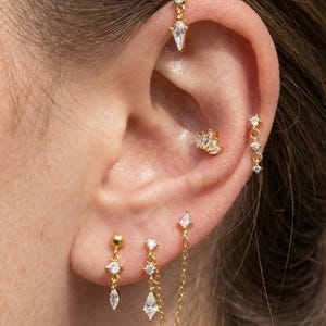 Könnte beinhalten: Nahaufnahme eines Ohrs, das mit mehreren goldenen Ohrringen geschmückt ist. Die Ohrringe weisen verschiedene Designs auf, darunter hängende tropfenförmige und rautenförmige Edelsteine. Ein Ohrring verbindet zwei Piercings mit einer filigranen Kette.