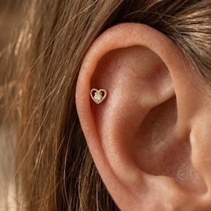 May include: A gold-colored heart-shaped earring with a small, clear gemstone in the center is shown in the ear. The earring is a stud style, and the ear is visible with the person's hair in the background.