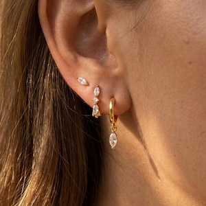 Può includere: Primo piano di un orecchio adornato con tre set di orecchini d'oro. Gli orecchini includono un piccolo borchia, un paio pendente e un orecchino a cerchio con una pietra a forma di goccia pendente. Gli orecchini sono incastonati con pietre chiare e scintillanti.