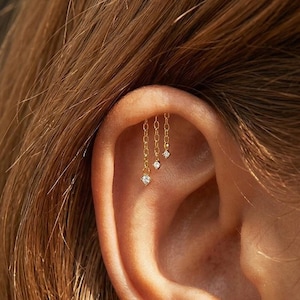 Könnte beinhalten: Goldene Kettenohrringe mit drei herabhängenden, diamantartigen Edelsteinen. Die Ohrringe sind am Ohrknorpel befestigt. Der Schmuck ist ein zartes und elegantes Accessoire.