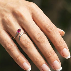 May include: A silver-toned bypass ring featuring a single pear-shaped, deep pinkish-red gemstone set at one end of the open band. The metal band has a smooth, polished finish and curves around the finger, leaving a gap between the two ends.