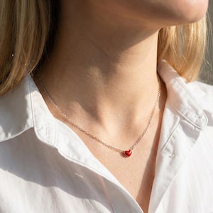 Puede incluir: Un delicado collar de plata con un pequeño colgante en forma de corazón rojo vibrante. El corazón está centrado en una fina cadena, creando un diseño simple pero elegante. El collar se lleva sobre una camisa blanca.