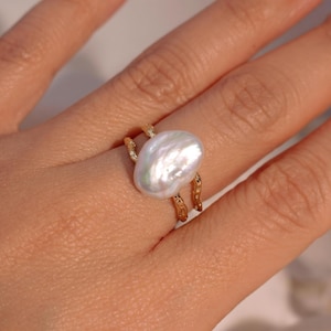 Op de afbeelding: Een goudkleurige ring met een grote, onregelmatig gevormde witte parel. De ringband heeft een textuur, organisch ontwerp, waarbij de parel prominent aanwezig is. De parel heeft een glanzende, iriserende glans.