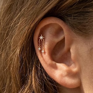 May include: A gold ear piercing featuring a starburst design at the top, with two dangling diamond-like charms. The earring is attached to the ear. The ear is in focus, with brown hair in the background.
