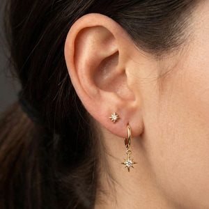 May include: An ear with two gold earrings. One is a small stud, and the other is a hoop with a dangling starburst charm. The earrings feature a small, sparkling stone at the centre. The earrings are gold in colour.