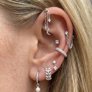 Puede incluir: Primer plano de una oreja adornada con múltiples pendientes de plata. Los pendientes incluyen un aro colgante con perla, un aro con forma de hoja, una cadena con un dije de luna y estrella, y un brazalete para la oreja con diamantes. La oreja está perforada en múltiples lugares.