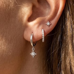 May include: A set of three silver-toned earrings consisting of a small starburst stud, a thin hoop encrusted with clear stones, and a larger smooth hoop featuring a dangling starburst charm with a central clear stone set in the middle.