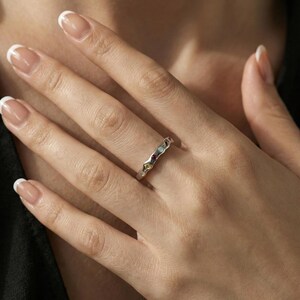 May include: A silver-toned ring featuring a wavy, organic band design. The ring is set with four small, round, multi-colored gemstones, including a light blue, a deep red, a yellow, and a clear stone, arranged along the center of the band.