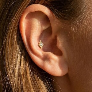 May include: A gold-coloured starburst earring with a small, clear gemstone in the centre is shown in an ear piercing. The earring is set against the backdrop of a person's ear and hair, with the focus on the jewellery.