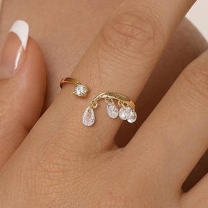 May include: A gold ring featuring a small, round, clear gemstone and a curved section with three hanging, teardrop-shaped clear gemstones. The ring is worn on a finger.