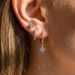 May include: A collection of gold earrings with blue gemstone accents. The set includes a small stud, a linear earring, and a hoop earring with a dangling floral charm. The earrings are displayed on an ear.