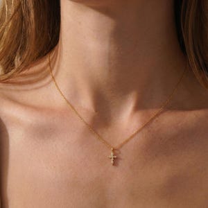 Pode incluir: Um delicado colar de ouro com um pequeno pingente de cruz adornado com pedras preciosas brilhantes. O pingente pende de uma corrente fina, criando um design simples, mas elegante.