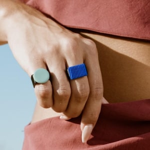 Può includere: Sono presentati due anelli. Uno ha una pietra circolare azzurra su una fascia nera. L'altro è un design rettangolare blu brillante. Lo sfondo è un cielo azzurro.
