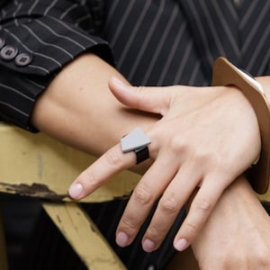 Könnte beinhalten: Ein geometrischer Ring mit einem grauen Dreieck und einem schwarzen Band wird am Finger getragen. Ein braunes und weißes quadratisches Armband befindet sich am Handgelenk. Die Person trägt einen schwarzen Nadelstreifenblazer.
