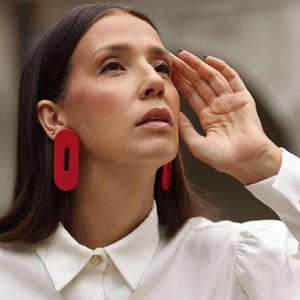 Pode incluir: Um par de brincos vermelho-brilhante em formato oval alongado com um recorte vertical central. Os brincos apresentam um acabamento liso e fosco e são usados nos lóbulos das orelhas, contrastando com uma camisa branca de colarinho.
