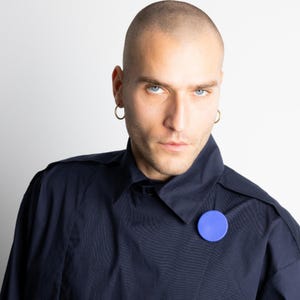 May include: A person wearing a navy blue jacket with a blue circular pin. The person has short hair and is wearing gold hoop earrings. The background is plain white.