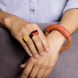 Può includere: Una mano con anelli e un braccialetto. Gli anelli presentano forme geometriche in giallo, arancione e bordeaux. Il braccialetto arancione ha una fascia intrecciata e la scritta "moonico".