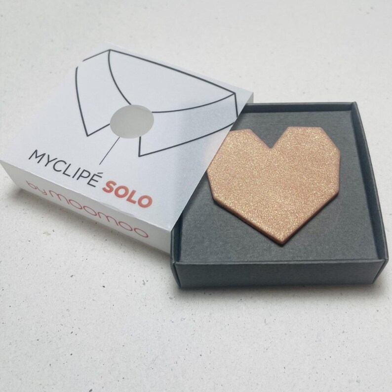 May include: A rose gold heart-shaped accessory, presented in a box. The box is white with a collar illustration and the text "MYCLIP&Eacute; SOLO" in black and red. The heart has a textured, glittery appearance.