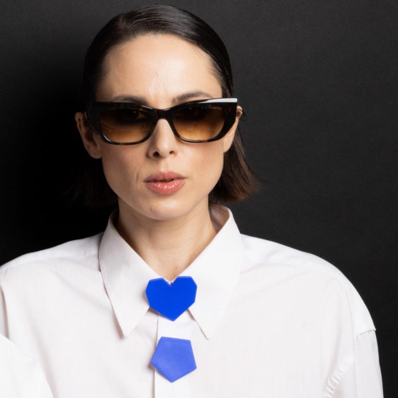 May include: A woman wearing dark sunglasses with a white-framed top. A blue heart-shaped and pentagon-shaped accessory is worn on the collar. The background is black.