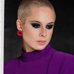 Peut inclure: Une personne aux cheveux très courts portant un haut violet à col montant et des boucles d'oreilles rouges circulaires. Le visage présente un maquillage des yeux charbonneux foncé et une expression neutre en regardant vers le bas.