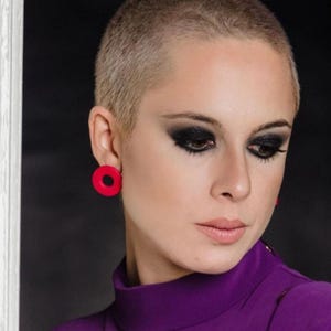 Könnte beinhalten: Eine Frau mit rasiertem Kopf trägt einen lila Rollkragenpullover und große rote Creolen.