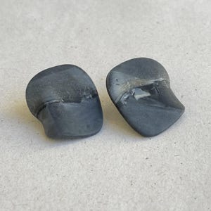 May include: Two dark gray, organic-shaped stud earrings. The earrings have a smooth, polished finish with a subtle, marbled effect. The design is minimalist and modern, suitable for everyday wear.