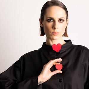 Peut inclure: Une chemise noire avec deux détails rouges en forme de cœur. Un cœur est placé au col, l'autre est tenu dans la main. La chemise a un col montant et des manches longues.