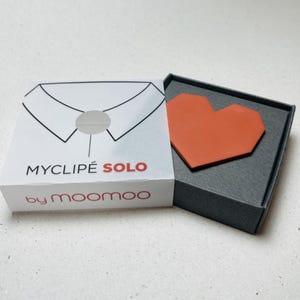 May include: A small, open gray box containing an orange heart-shaped object. The box is next to a white box with the text "MYCLIP&Eacute; SOLO" and "by moomoo" printed on it. The white box has a graphic of a shirt collar.