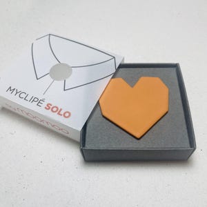 May include: An open gray box with an orange heart-shaped object inside. The box is next to a white box with the text "MYCLIP&Eacute; SOLO" and a diagram of a shirt collar. The brand name "moomoo" is also visible.