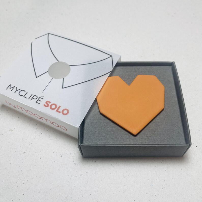 May include: An open gray box with an orange heart-shaped object inside. The box is next to a white box with the text "MYCLIP&Eacute; SOLO" and a diagram of a shirt collar. The brand name "moomoo" is also visible.