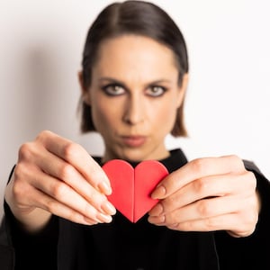 May include: A bright red heart shape held in front of a person. The heart is made of two connected pieces. The person is wearing a black top and has short dark hair. The background is plain white.