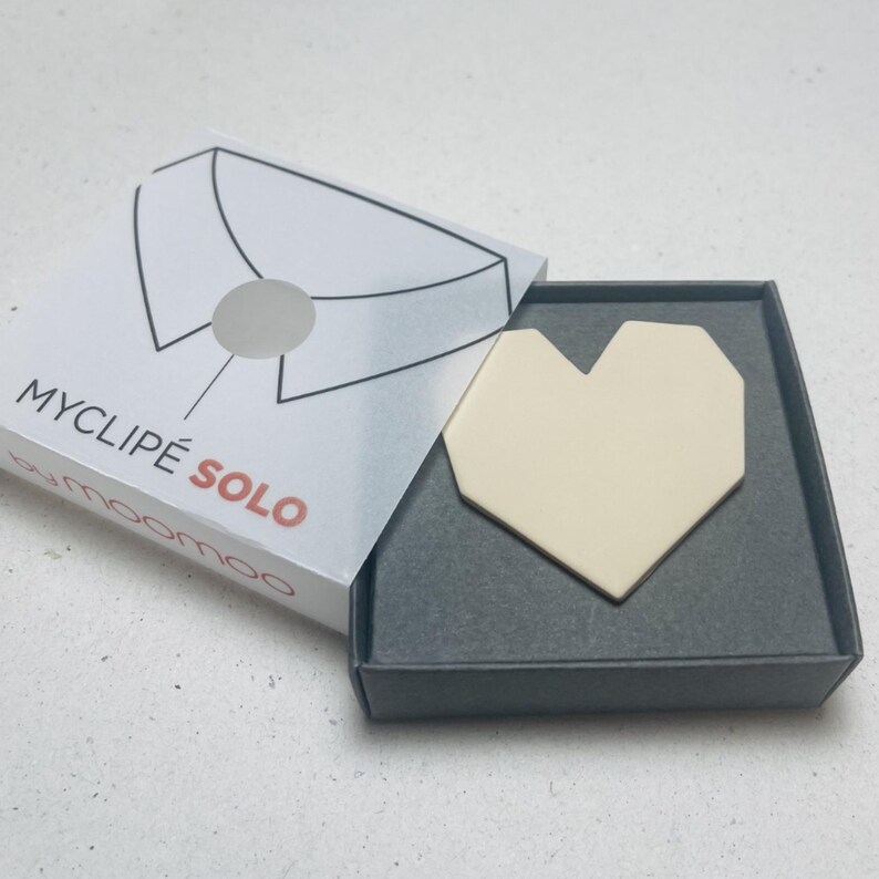 May include: A cream-colored heart-shaped accessory is displayed inside a gray box. The box is open, revealing the product and its packaging. The packaging features a collar illustration and the text "MYCLIP&Eacute; SOLO".