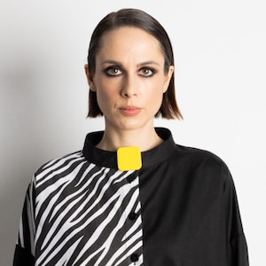 May include: A black and white zebra print shirt with a black collar and a bright yellow square clasp. The shirt is buttoned down the front and has long sleeves. The person in the image has short, dark hair and is looking directly at the camera.