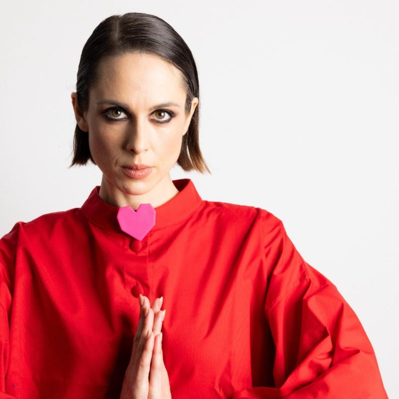 May include: A red long-sleeve shirt with a high collar and a pink heart-shaped brooch. The person is wearing the shirt and has their hands together in a prayer position. The background is white.