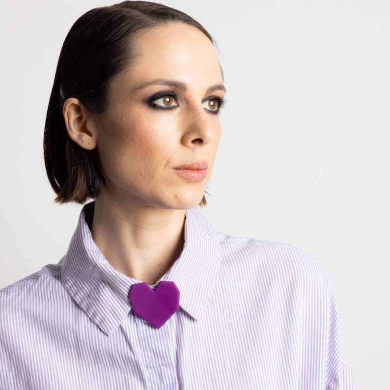May include: A purple heart-shaped brooch pinned to a striped button-down shirt. The brooch is a solid, vibrant purple color and is positioned at the collar. The shirt has thin, vertical stripes in a light purple hue.