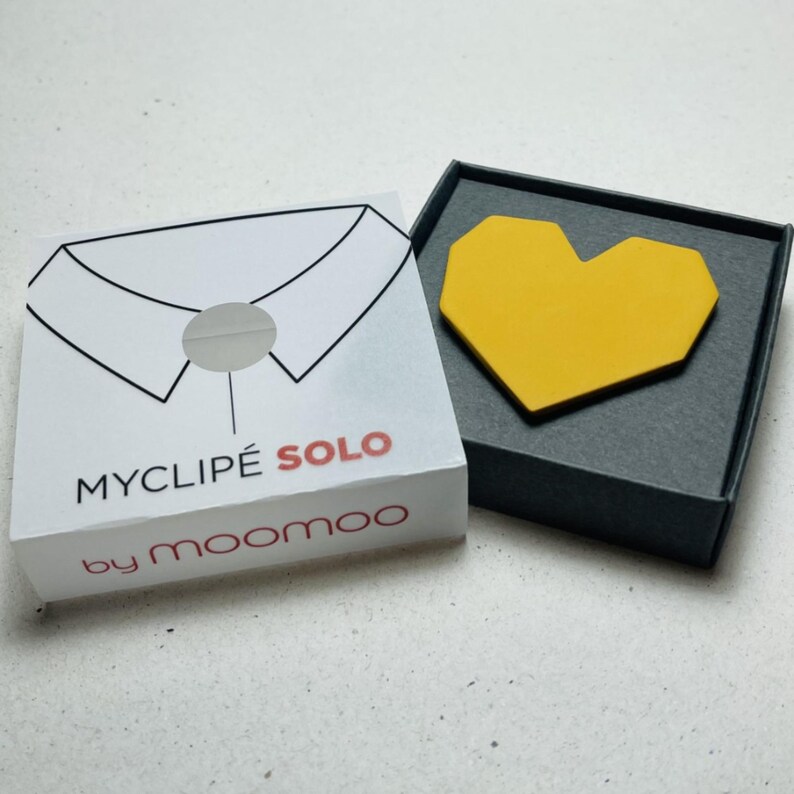 May include: A yellow heart-shaped accessory sits in a dark gray box next to a white box. The white box has a drawing of a shirt collar and the text "MYCLIP&Eacute; SOLO by moomoo". The heart-shaped accessory is made of a solid yellow material.