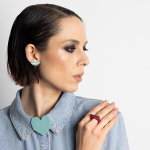May include: A woman wearing geometric jewelry, including a teal heart-shaped brooch, matching earrings, and a red and blue ring. She is wearing a denim shirt. The jewelry has a modern, minimalist design.