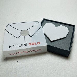 May include: A white box with the text "MYCLIP&Eacute; SOLO" and "by moomoo" in red and black. The box has a collar design. Next to the box is a dark gray box with a white heart-shaped object inside.