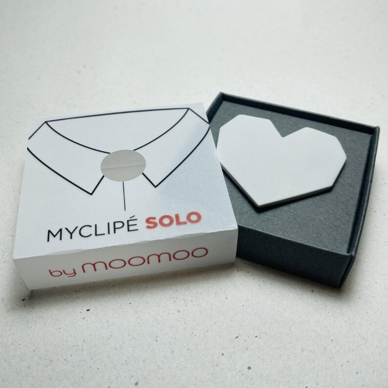 May include: A white box with the text "MYCLIP&Eacute; SOLO" and "by moomoo" in red and black. The box has a collar design. Next to the box is a dark gray box with a white heart-shaped object inside.