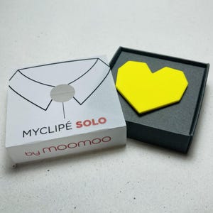 May include: A white box with a collar illustration and the text "MYCLIPE SOLO by moomoo" next to a black box containing a yellow heart-shaped object. The boxes are on a light surface.