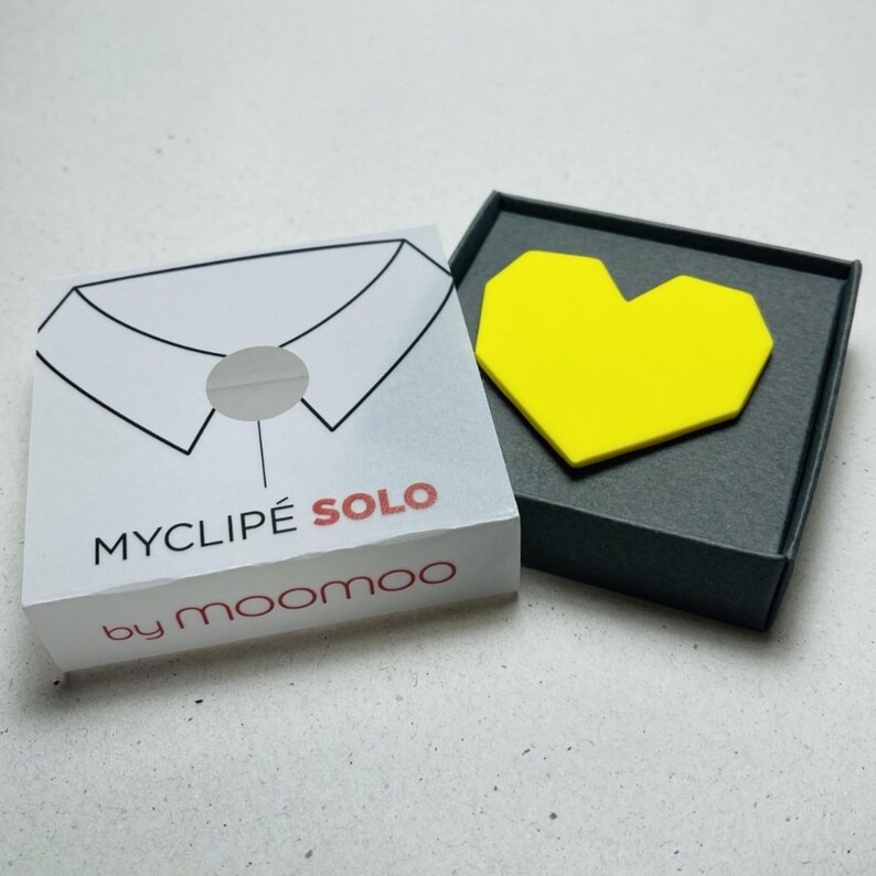May include: A white box with a collar illustration and the text "MYCLIPE SOLO by moomoo" next to a black box containing a yellow heart-shaped object. The boxes are on a light surface.