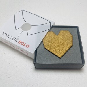May include: A gold-colored heart-shaped lapel pin in a gray box. The pin has a textured surface and is displayed with its packaging, which features the text "MYCLIP&Eacute; SOLO". The box is open, revealing the pin.
