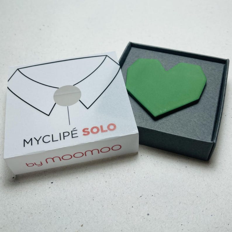 May include: A white box with the text "MYCLIPE SOLO" and "by moomoo" features a diagram of a shirt collar. A green heart-shaped object rests in an open gray box. The items are on a white surface.