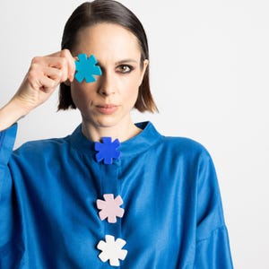 May include: A woman wearing a blue top with three flower-shaped buttons in blue, pink, and white. She holds a turquoise flower-shaped object over one eye. The background is plain white.