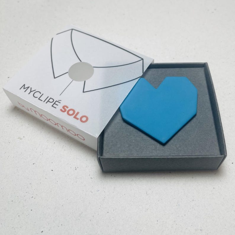 May include: A turquoise heart-shaped accessory rests in a gray box. The box is open, revealing the product and its packaging. The packaging features a white box with a shirt collar design and the text "MYCLIPE SOLO" in black and red.